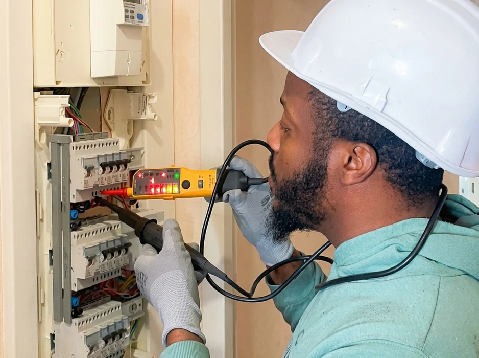 Électricien en intérieur, casque blanc et gants de protection, analyse un tableau électrique