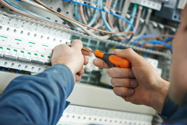 Artisan en chemise à carreaux tenant un multimètre numérique jaune, portant une ceinture à outils professionnelle grise et orange équipée de tournevis, pinces et instruments de mesure électrique