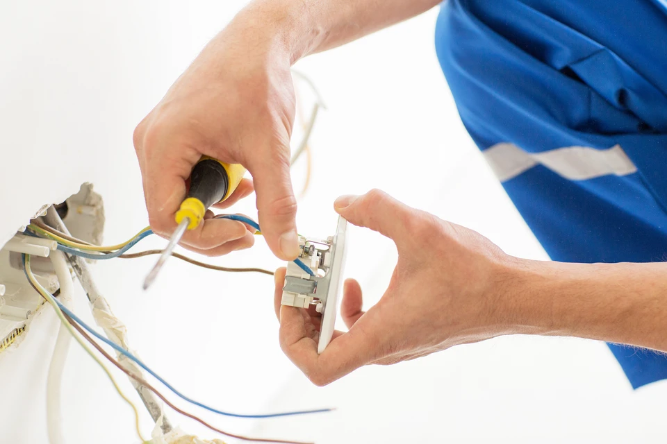 Électricien en bleu travaillant sur installation électrique murale, manipulant câbles et composants avec tournevis