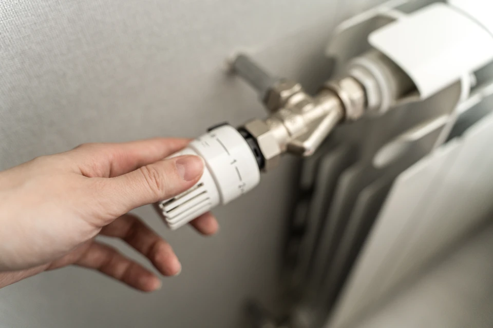 Homme en chemise à carreaux effectuant des travaux de plomberie sur un radiateur blanc dans un intérieur résidentiel lumineux près d'une fenêtre