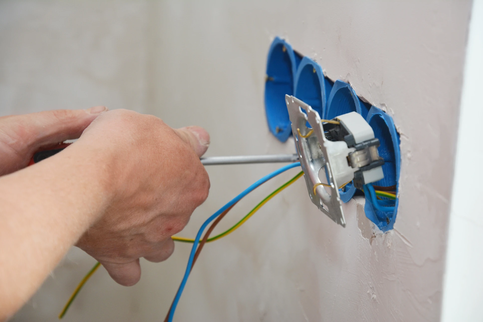Électricien en tenue de travail bleue et blanche avec casque de sécurité blanc, agenouillé devant un mur blanc, travaillant sur une installation électrique près d'une prise murale
