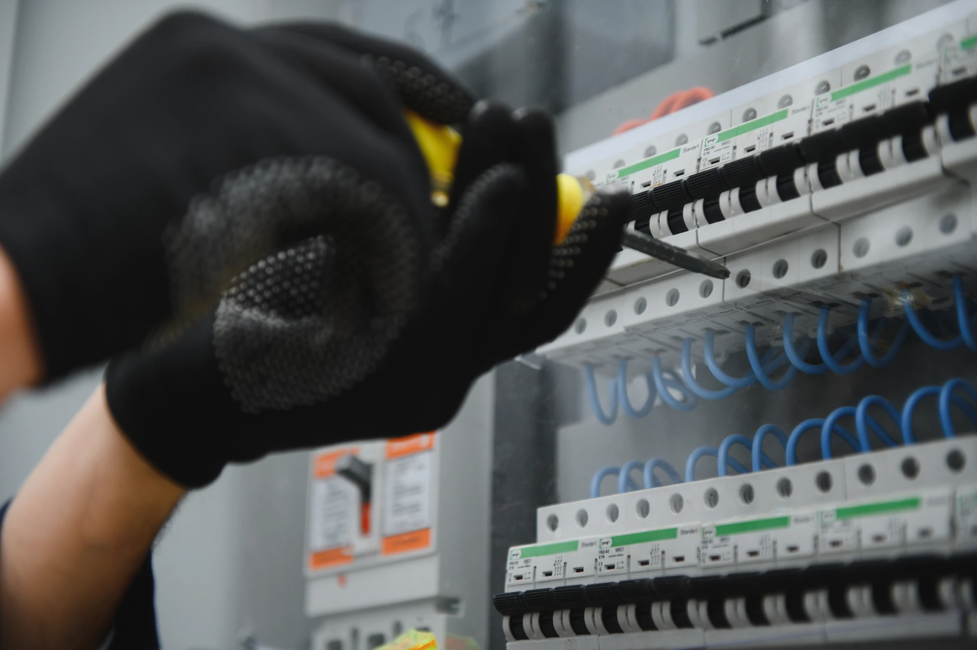 Photographie d'un électricien artisan en vêtements de travail professionnels réalisant l'installation d'une pompe à chaleur ou d'un système électrique dans l'intérieur d'une maison résidentielle. Le professionnel est concentré sur son travail, entouré de matériel et d'outils spécialisés (équipements de chauffage, panneaux électriques, câbles). La lumière naturelle illumine la scène. L'atmosphère doit être professionnelle, sérieuse et refléter l'expertise artisanale et la qualité d'exécution.